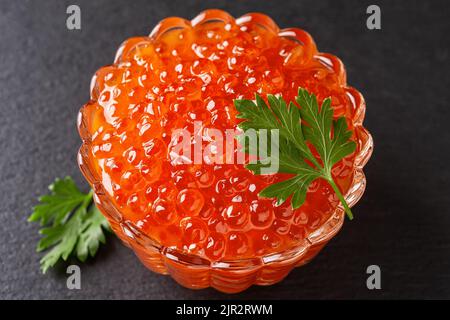 Salmon red caviar in a glass bowl on a black background. Crystal dish full of trout caviar close-up. Seafood fish delicacy of salted salmon roe. Stock Photo