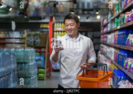 Experienced mature Asian shopper in supermarket, man smiling and reading message on smartphone, makes purchases and chooses products from various shelves Stock Photo