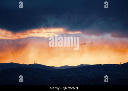 Dramatic sunset storm clouds with rainfall over rolling hills and distant wind turbines Stock Photo