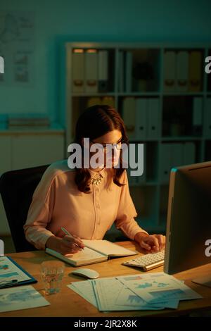 Young woman reading data on computer screen and taking notes Stock Photo
