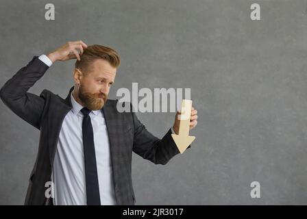Confused businessman in suit holding arrow down scratching head studio shot Stock Photo