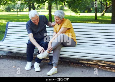 During morning stroll, sportive exercises in park, elderly 60s woman got injured ankle, grip foot sit in bench with caring disappointed husband. Traum Stock Photo