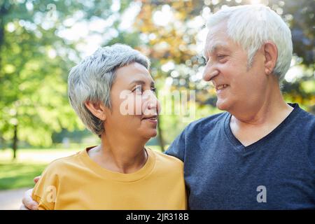 Pretty multi racial elderly 60s couple stroll walk in park smiling looking at each other with tenderness and love. Happy endless marriage, romantic re Stock Photo