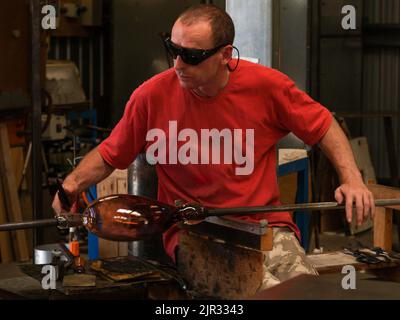 Glassblower with dark protective eye glasses and with molten glass shape on long pole turning and shaping it Stock Photo