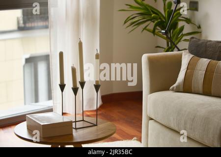 corner of a living room with a bay window, a round wooden side table with a chandelier and a sofa with cushions Stock Photo
