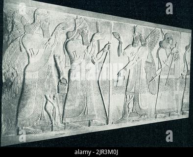 The 1910 caption reads: 'Assyrian king between genie - in the Royal Museum in Berlin. The Assyrian mythical guardian figure, called a Genius, wears a two-horned helmet and fringed garment and carries a double-handled dagger, all attributes of his divine nature. His hands gesture protectively toward a flowering tree, just visible at the right edge of the sculpture. This relief, once brightly painted, is a fragment from the Northwest palace built by the Assyrian king Ashirnasirpal II (883—859 BCE) in his capital city at Kalhu, now the Iraqi city of Nimrud. Dozens of huge stone slabs carved with Stock Photo