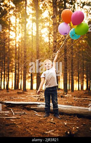 Balloons make me excited. a happy little boy holding a bunch of balloons while standing outside in the woods. Stock Photo