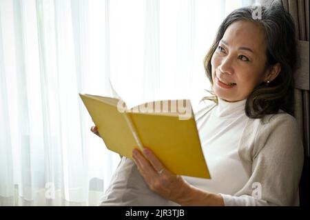 A charming and peaceful Asian middle-aged woman reading her favorite book in the living room. Lifestyle concept. Stock Photo