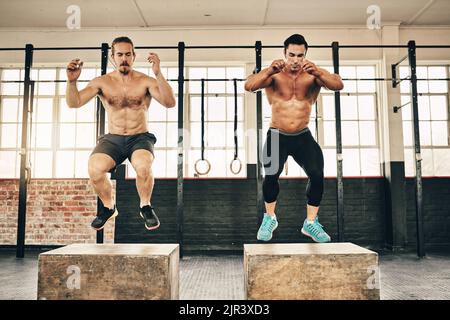 Jumping just got a lot more tougher. two fit and determined young men jumping and doing squats to workout in a gym. Stock Photo