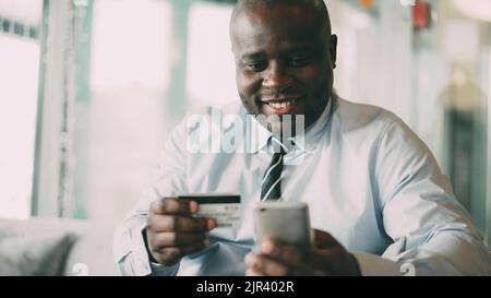 Happy African American businessman in formal clothes smiling and paying online bill while keeping credit card and smartphone in his hands in glassy cafe Stock Photo