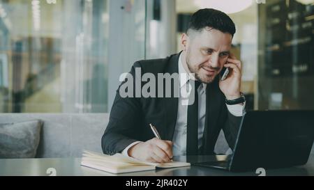 Portrait of cheerful Caucasian businessman sitting, smiling, talking on smartphone with his friend and writing down information in notepad while looking at laptop in modern cafe during lunch time Stock Photo