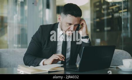 Smart Caucasian businessman sitting, talking on smartphone and writing down business information in his notepad while looking at laptop in glassy cafe Stock Photo