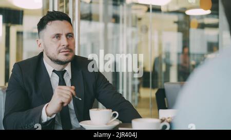 Bearded Caucasian entrepreneur gesticulating and discussing his startup plan with investor in formal wear in glassy cafe during during meeting Stock Photo