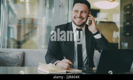Happy bearded businessman in black business suit sitting at the table, smiling and talking on mobile phone in modern cafe. Man keeps a notebook at his hand Stock Photo