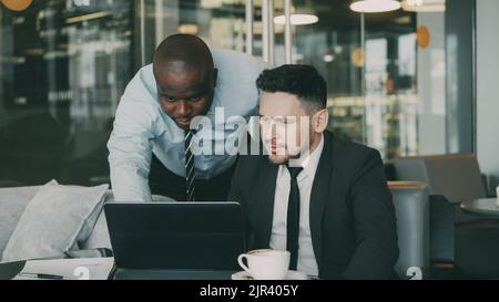 Two business partners discussing actively their startup project sitting in stylish cafe. Bearded Caucasian businessman sitting ang talking African American colleague while looking at laptop computer Stock Photo