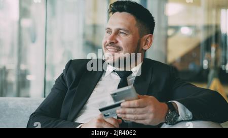 Cheerful Caucasian businessman in suit using online banking holding credit card and smartphone in his hands in modern cafe indoors during lunch break Stock Photo