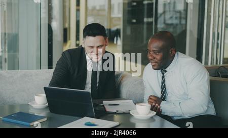 Two multi ethnic businessmen looking at laptop computer and laughing in glassy cafe. Men having fun and joking. African American businessman and Caucasian man feel success. Stock Photo
