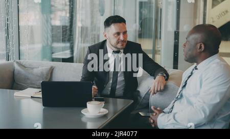 Two ambitious entrepreneurs brainstorming business ideas of their startup in modern cafe. African American man in white shirt puts down something and bearded Caucasian businessman keeps a tablet Stock Photo
