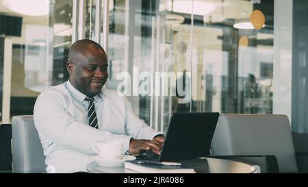 Portrait of hilarious African American businessman in formal clothes smiling, printing and working on his laptop in airy cafe during lunch break. Stock Photo
