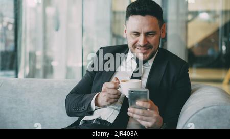 Smiling bearded Caucasian businessman in formal clothes smiling, relaxing and looking at his smartphone screen while drinking coffee in airy cafe during lunch time Stock Photo