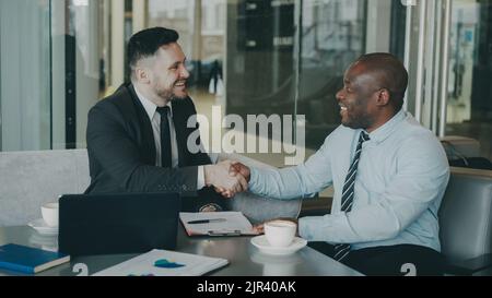 Two business colleagues discussing their business contract and looking at notepad in stylish cafe indoors. African American businessman puts his signature and shakes the hand of Caucasian man Stock Photo
