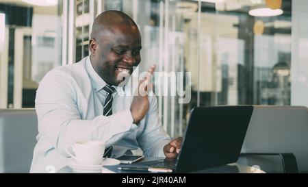 African Americanbusinessman in formal wear having online video call with his laptop and talking to his family in classy cafe during lunch break. He smiles and waving his hand cheerfully. Stock Photo