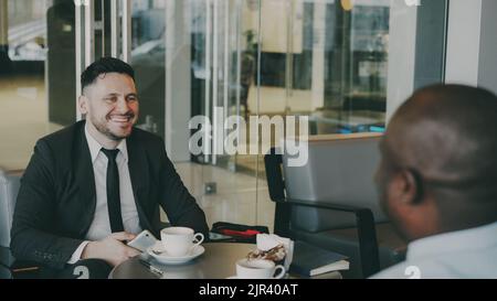 Two multi-ethnic businessmen sitting, smiling and discussing their startup in airy cafe during lunch time. Cheery African American partner strokes his knees happily. Coffee cups are on their table Stock Photo