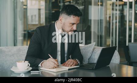 Smart Caucasian businessman using laptop computer and writing down information in his notepad in glassy cafe having coffee cup on his table. A cup of coffee is on his table. Stock Photo