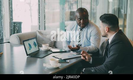 Two confident businessmen looking at diagrams and graphs on laptop computer and discussing the financial report and statistics of their business project in modern glassy cafe during lunch time Stock Photo