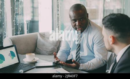 African and Caucasian businessmen looking charts and diagrams on laptop computer and discussing financial aspects of start-up project in cafe during coffee break Stock Photo