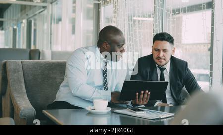 Cheerful African American businessman in formal clothes using digital tablet discussing startup ideas with his caucasian partner in stylish cafe indoors during lunch break Stock Photo