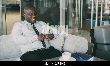 Cheerful African American businessman in formal clothes using smartphone in cafe. He looks at screen and smiles happily, with a cup of coffee on his table. Stock Photo