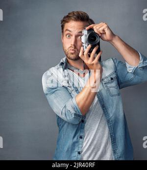 Keep calm and snap on. Portrait of a happy young man holding up a camera while posing against a gray background in the studio. Stock Photo