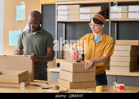 Multiethnic couple of workers packing parcels in cardboard boxes together before shipping Stock Photo