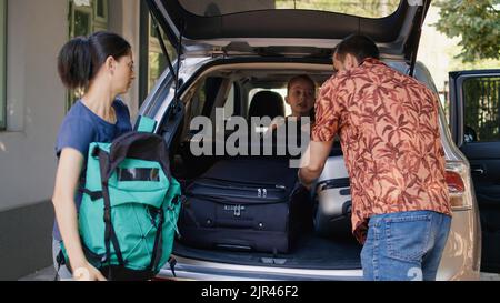 Family getting ready for summer field trip while loading vehicle with voyage trolleys and citybreak. Parents putting luggage in car trunk while unpatient daughter hurrying them. Stock Photo