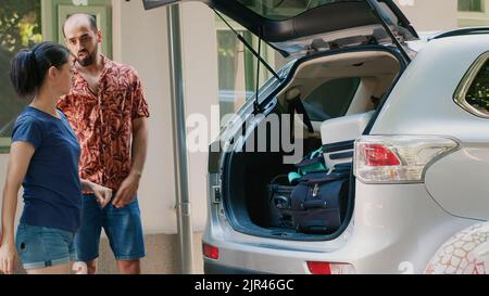 Caucasian family loading voyage luggage inside car trunk while going on summer holiday field trip together. People getting ready for weekend citybreak while putting baggage inside vehicle. Stock Photo