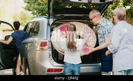 Mother, daughter and grandparents putting baggage and trolleys inside vehicle while going on field trip. Family loading voyage luggage in car trunk while getting ready for summer holiday citybreak. Stock Photo