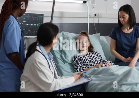 Pediatrist examining sick little girl disease symptoms in children healthcare facility room while mother sitting beside her. Ill daughter under treatment getting consulted by hospital medical staff. Stock Photo