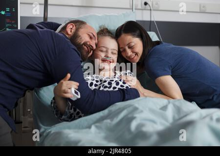 Happy sick little girl hugged by joyful smiling parents in hospital pediatrics ward. Cheerful mother and father hugging ill daughter sitting in patient bed while under treatment. Stock Photo