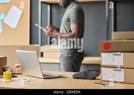 Horizontal image of workplace with laptop and cardboard boxes on it with man working in background in warehouse Stock Photo