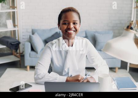 Confident African-American businesswoman looking and smiling at the camera, using laptop in the office writing notes in diary. Multiracial female Stock Photo