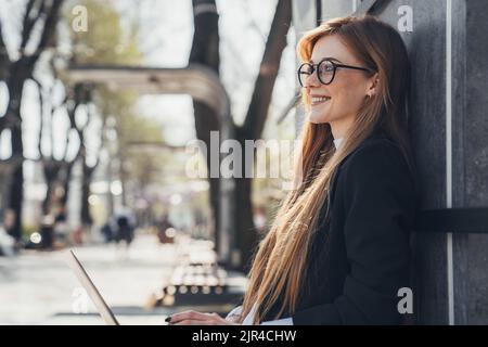 Red-haired casual woman with freckles is resting on a bench in the city park, on a summer day, using laptop. People urban lifestyle concept Stock Photo