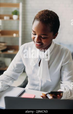 African businesswoman with laptop writing something in notebook while using the mobile phone. Working on the project, typing email on the laptop Stock Photo
