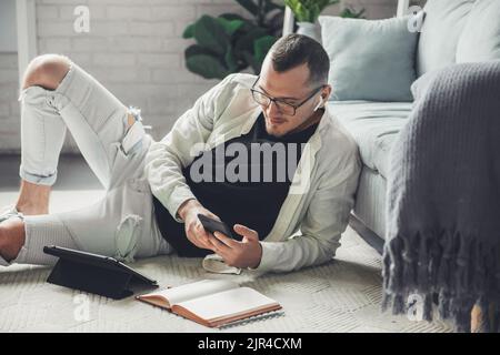 Man in eyeglasses with mobile phone studying from home lying on the floor in a modern and stylish interior. Education internet technology. Distance Stock Photo