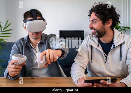 Caucasian men enjoying virtual reality glasses while sitting at the table in home. Two happy guys gesturing and using vr headset or 3d spectacles play Stock Photo