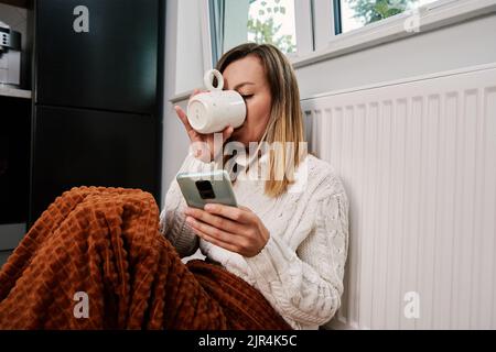 Worried sad woman sits under blanket near heating radiator and use smartphone, Rising costs in private households for gas bill due to inflation and wa Stock Photo