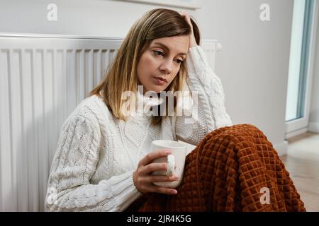 Worried sad woman sits under blanket near heating radiator with cup of tea, Rising costs in private households for gas bill due to inflation and war, Stock Photo
