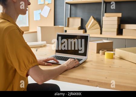 Close-up of warehouse worker sitting at table and typing on laptop to work with online orders in distribution centre Stock Photo