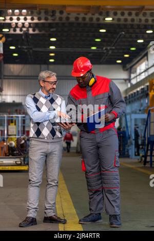 Multiracial Industrial Co-Workers Standing In A Factory And Talking About Production Process. African-American Worker Writing On Clipboard. Stock Photo