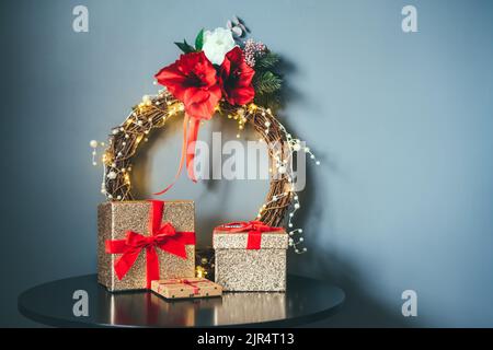Few glittering golden gift boxes with velvet red ribbon and elegant Christmas flower wreath on the black table on gray wall background. Minimalist fes Stock Photo